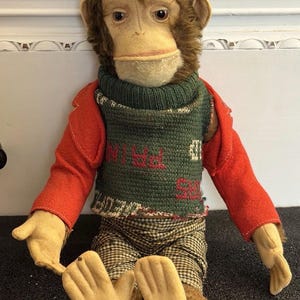 May include: A plush monkey doll with brown fur, a beige face, and hands. It wears a red jacket over a green knitted sweater with red text, and brown and white checkered pants. The doll is in a seated position with its arms extended outward.