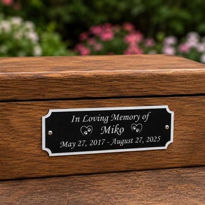Könnte beinhalten: Eine rechteckige Holzbox mit dunkelbrauner Oberfläche und Klappdeckel. Auf der Vorderseite befindet sich ein schwarzes Metallschild mit der Gravur: In Loving Memory of Miko, May 27, 2017 - August 27, 2025, sowie zwei Herz-Pfotenabdruck-Symbolen.