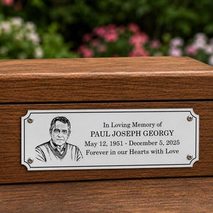 Puede incluir: Una urna funeraria rectangular de madera con acabado de veta marrón oscuro. Una placa de metal plateado está fijada al frente, con un retrato en tinta negra de un hombre y el texto: In Loving Memory of PAUL JOSEPH GEORGY May 12, 1951 - December 5, 2025 Forever in our Hearts with Love.