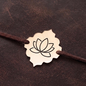 May include: A gold-toned metal charm with a scalloped edge and a black engraved lotus flower design, threaded onto a thin dark brown leather cord. The charm is centred on a dark brown textured surface.