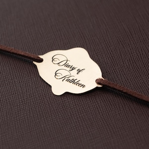 May include: A dark brown textured cover features a gold-colored metal plaque with a scalloped edge. The plaque is engraved with the script text "Diary of Kathleen" and is attached to a thin, dark brown cord that runs horizontally across the cover.