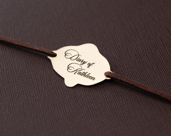 May include: A dark brown textured cover features a gold-colored metal plaque with a scalloped edge. The plaque is engraved with the script text "Diary of Kathleen" and is attached to a thin, dark brown cord that runs horizontally across the cover.