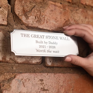 Peut inclure: Une plaque rectangulaire en métal argenté aux coins décoratifs portant le texte gravé : THE GREAT STONE WALL, Built by Daddy, 2021-2026, Worth the wait. La plaque présente une surface lisse et réfléchissante avec une fine bordure sombre.