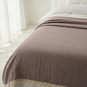 May include: A mauve-colored blanket with a textured weave is draped over a bed. The blanket has a fringe trim along the bottom edge. White sheets and pillows are visible beneath the blanket. The scene is lit by natural light.