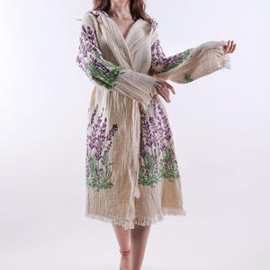 Peut inclure: Une robe crème à manches longues avec un design enveloppant. La robe présente des motifs floraux brodés de lavande et de vert sur les manches et la jupe. L'ourlet a une bordure effilochée.