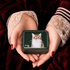 May include: A dark green octagonal box featuring a painted portrait of a ginger cat on the lid with the name Holly written in gold script. The side of the box is engraved with the text James & Grace and the date 08-20-2014 in gold lettering.