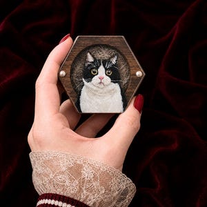 以下が含まれることがあります： 黄色い目とピンクの鼻を持つ白黒の猫のニードルフェルトの肖像画が入った六角形のダークウッドフレーム。猫は質感のあるベージュの背景の前に配置されており、フレームには2つの小さな白いパールビーズがアクセントとしてあしらわれています。