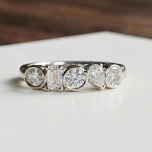 May include: A silver-toned ring featuring five bezel-set clear stones in various shapes, including round, emerald, and pear cuts, arranged in a horizontal row along the band. The metal has a polished finish and a smooth, continuous circular band.