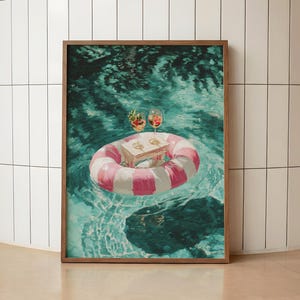 May include: A framed art print depicting a pink and white striped inflatable pool float drifting on deep turquoise water. A small wooden tray sits in the centre of the float, holding two stemmed glasses filled with a reddish beverage and garnish.