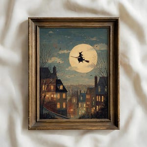 May include: A framed wall art print depicting a silhouette of a witch flying on a broomstick against a large, glowing full moon. The scene features a town with multi-story buildings, lit windows, and bare trees under a night sky with clouds.
