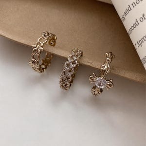 May include: Three gold-toned rings arranged in a row. The first ring features a repeating openwork floral pattern. The second ring has a lattice design with embedded clear stones. The third ring displays a central cross motif with a clear stone.
