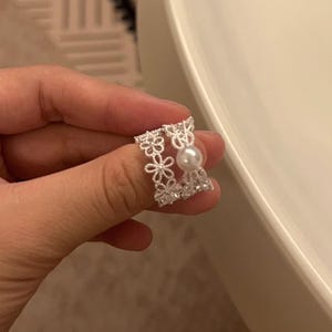 Puede incluir: Un anillo de tono plateado con un diseño calado de pequeñas flores conectadas y una cuenta esférica blanca. La banda está construida con filigrana de metal delicada, creando una apariencia de encaje con un acabado pulido y brillante.