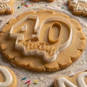 Peut inclure: Un emporte-pièce en plastique blanc en forme de chiffre 40 avec le mot forty en écriture cursive à l'intérieur, pressé dans un morceau de pâte à biscuit ronde beige clair. L'outil présente un chiffre 4 gras et un zéro avec le texte forty.