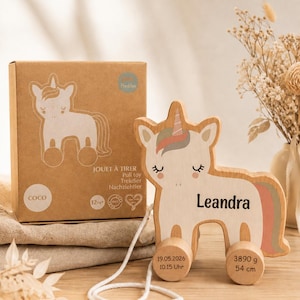 Könnte beinhalten: Ein hölzernes Einhorn-Nachziehtier mit weißem Körper, rosa-grauer Mähne und Horn. Der Name Leandra steht auf der Seite. Die Räder sind mit 19.05.2026, 10.15 Uhr, 3890 g und 54 cm bedruckt. Es steht neben einer braunen Pappschachtel mit Aufdruck.
