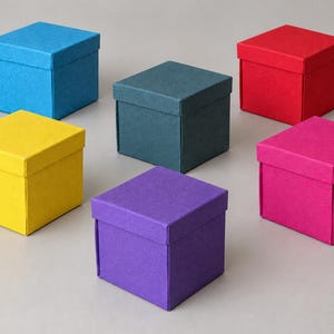 Könnte beinhalten: Eine Ansammlung von sechs quadratischen Geschenkboxen mit Deckeln, jede in einer anderen kräftigen Farbe wie leuchtendes Blau, dunkles Petrol, kräftiges Rot, sonniges Gelb, sattes Violett und Pink, die in einer Gruppe angeordnet sind.