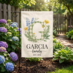 May include: A rectangular garden flag featuring a large green and white gingham letter G surrounded by blue and yellow floral illustrations. The text reads GARCIA family EST. 2018 in black lettering. The flag hangs from a black metal stand.