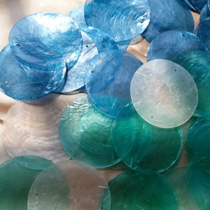Peut inclure: Une collection de disques en coquillage plats et circulaires dans diverses nuances de bleu, de sarcelle et de blanc translucide. Chaque disque présente une surface lisse et irisée avec un éclat naturel et deux petits trous pré-percés.