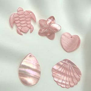 Puede incluir: Una colección de cinco colgantes de concha de color rosa claro iridiscente con varias formas, incluyendo una tortuga marina, una flor de cinco pétalos, un corazón, una lágrima y una concha festoneada, cada uno con un pequeño orificio.