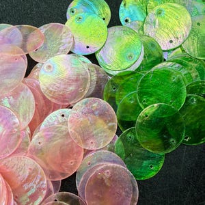 May include: A collection of circular, flat shell sequins featuring an iridescent finish. The sequins are arranged in two distinct colour groups, with one side displaying soft pink and peach tones and the other side showing vibrant green hues.