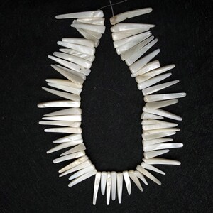 May include: A strand of white mother of pearl beads shaped like elongated, tapered spikes or teeth. The beads are arranged in a circular formation, showcasing a smooth, iridescent surface with natural variations in the off-white colour tones.