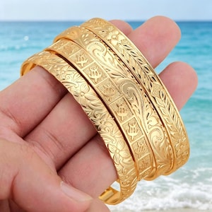 May include: Three gold-toned metal bangles featuring intricate embossed patterns. The bracelets display a combination of floral motifs, scrolling leaf designs, and a repeating grid pattern containing small turtle-shaped engravings.