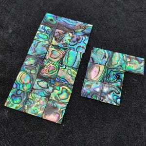 May include: A collection of small, square abalone shell tiles arranged in two groups. Each tile features a unique, iridescent pattern with swirling colours of deep blue, vibrant green, purple, and pink, reflecting light with a natural pearlescent sheen.