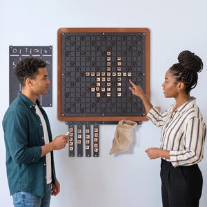 Könnte beinhalten: Ein wandmontiertes Wortspielbrett mit einem dunklen Gitter und hölzernen Buchstabenplättchen, die ein Kreuzworträtsel bilden. Unter dem Brett befinden sich vier vertikale Leisten für Buchstaben und ein kleiner beiger Stoffbeutel.