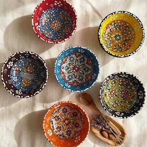 May include: Five ceramic bowls with intricate floral patterns in red, yellow, dark brown, blue, and orange, each featuring a white dotted rim. A small wooden dish holds a wooden spoon, two almonds, and a sprig of dried lavender nearby.