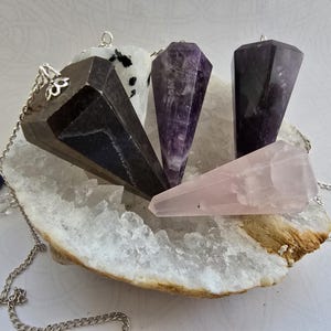 May include: Four faceted gemstone pendulums in dark gray, purple, deep violet, and pale pink, each attached to a silver-toned metal chain. The pendulums are arranged on a large, rough-edged white crystal geode cluster with visible internal points.