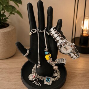 Pode incluir: Um porta-joias preto em forma de mão exibindo vários acessórios. Um relógio prateado com mostrador branco e uma pulseira prateada estão nos dedos. Colares, pulseiras e anéis de prata com berloques também são exibidos. O suporte fica em uma base redonda.