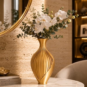 Pode incluir: Um vaso de metal dourado com textura canelada vertical e gargalo estreito, contendo um buquê de flores de orquídea brancas e caules de eucalipto verdes. O vaso tem um corpo arredondado que se estreita em direção à base e à abertura.