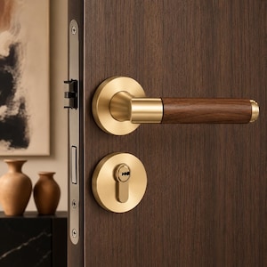 Peut inclure: Une poignée de porte composée d'un levier horizontal avec une prise en bois brun foncé et une base en métal doré brossé. L'ensemble comprend une plaque de serrure circulaire assortie et une têtière métallique verticale sur une porte en bois.