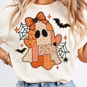 May include: A cream-colored t-shirt featuring a central graphic of a ghost made of various orange, white, and brown patchwork patterns. The ghost wears a plaid bow and is surrounded by black spiderwebs, spiders, bats, and orange star shapes.