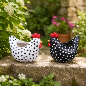 May include: Two chicken-shaped planters, one white with black polka dots and one black with white polka dots. Both feature a bright red comb, a small orange beak, and a hollow center for plants, resting on a stone surface in a garden setting.