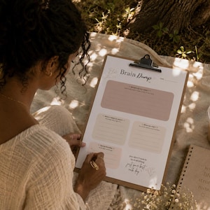 Puede incluir: Un portapapeles con una hoja de trabajo "Brain Dump", con indicaciones y espacio para notas. La hoja de trabajo tiene una paleta de colores neutros con acentos florales. También se ven un cuaderno, café helado y un bolígrafo.