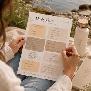 Może przedstawiać: Planer "Daily Reset" z kwiatowymi akcentami, zawierający sekcje na intencje, priorytety i zadania. Widoczna jest również biała butelka na wodę z drewnianą pokrywką i napisem "soft reset co.". Do pisania użyto złotego długopisu.