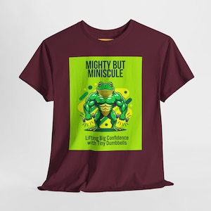 Peut inclure: T-shirt bordeaux avec une grenouille de dessin animé musclée tenant de petits haltères. La grenouille est verte avec des accents jaunes, sur un fond vert citron. Le texte indique "MIGHTY BUT MINISCULE" et "Lifting Big Confidence with Tiny Dumbbells".