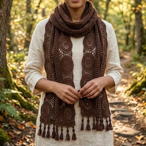 May include: A dark brown crochet scarf featuring a repeating circular motif pattern with an openwork grid design. The scarf is finished with a series of small, dangling tassels along the bottom edge, providing a textured and detailed appearance.