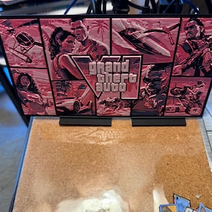 May include: A rectangular display sign featuring a pink-toned collage of Grand Theft Auto VI imagery, including characters, vehicles, and palm trees. The central logo reads Grand Theft Auto VI in a bold, stylized font. The sign is mounted on two black stands.