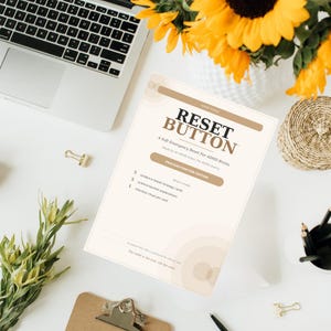 Puede incluir: Un documento blanco titulado "RESET BUTTON" con el texto "A Soft Emergency Reset For ADHD Brains". El documento está rodeado por un portátil, girasoles y material de oficina.