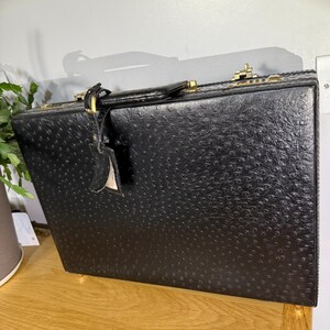 May include: A black briefcase with a textured ostrich-skin pattern, featuring a top handle, gold-tone metal hardware, and a matching luggage tag attached to the handle. The rectangular case includes secure locking mechanisms on the top edge.