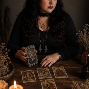 May include: A deck of tarot cards with dark, ornate covers held in one hand, with four tarot cards laid out on a wooden surface. The cards feature detailed illustrations and text. Two lit candles in metal holders are placed in the foreground.