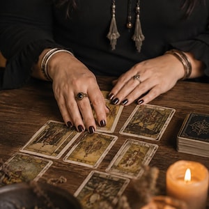 May include: A set of rectangular tarot cards with vintage-style illustrations laid out on a wooden surface. The cards feature muted tones and intricate artwork. Two lit candles in glass holders and a dark bowl are positioned near the cards.