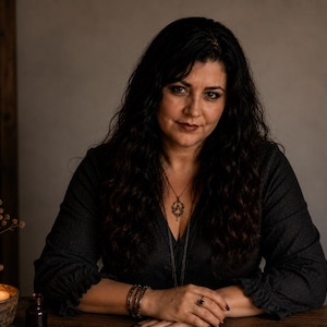 May include: A person with long dark wavy hair wearing a dark charcoal long-sleeved top with ruffled cuffs, a gold pendant necklace, and stacked bracelets. The person has dark lipstick and is positioned behind a wooden surface with a lit candle.