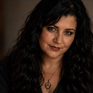May include: A person with long, dark, wavy hair wearing a dark-colored top and a silver-toned necklace featuring a circular pendant with a detailed, intricate design. The person has a neutral expression and is looking directly toward the camera.