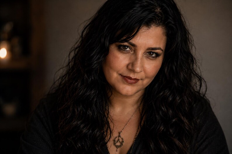 May include: A person with long, dark, wavy hair wearing a dark-colored top and a silver-toned necklace featuring a circular pendant with a detailed, intricate design. The person has a neutral expression and is looking directly toward the camera.