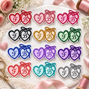 May include: A collection of twelve heart-shaped patches arranged in four rows. Each patch features two connected hearts with a bow on top, displaying the letters F and B in a decorative font. The patches come in various colors including red, pink, black, gold, green, blue, purple, teal, and gray.