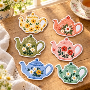 May include: Six embroidered teapot-shaped patches featuring floral designs. The collection includes a yellow, coral, olive green, pink, blue, and light green teapot, each decorated with white, yellow, or red flower motifs and green leaves.