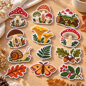 May include: A collection of embroidered patches featuring various forest-themed designs including red and white spotted mushrooms, yellow chanterelles, a brown snail, a green fern leaf, an orange oak leaf, a moth, acorns on an oak branch, and red berries.