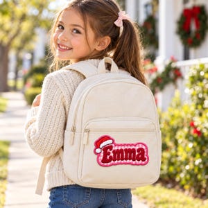 K&ouml;nnte beinhalten: Ein cremefarbener Rucksack aus Segeltuch mit einem personalisierten Aufn&auml;her, der den Namen Emma in roten Buchstaben zeigt, umrandet von einem rosa-wei&szlig; karierten Muster und verziert mit einer kleinen rot-wei&szlig;en Weihnachtsm&uuml;tze.